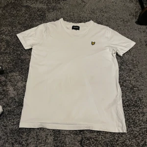 Vit t-shirt från Lyle & Scott - Säljer en stilren vit t-shirt från Lyle & Scott med ett broderat emblem på bröstet. T-shirten har en klassisk rund hals och korta ärmar. Perfekt för en avslappnad look.