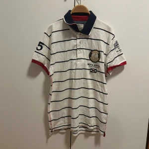 Vit och marinblå randig pikétröja från Selected Homme - Säljer en stilren vit pikétröja med marinblå ränder från Selected Homme. Tröjan har korta ärmar med röda detaljer och en klassisk krage. Perfekt för en avslappnad men ändå snygg look.