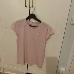 Rosa t-shirt från Tommy Hilfiger - Säljer en söt rosa t-shirt från Tommy Hilfiger med volangdetaljer på ärmarna. Den har en liten logga på bröstet och är i en mjuk bomullskvalitet. Perfekt för en avslappnad stil. Fin rosa färg nu t våren/sommaren säljer pgv används nt så mycket, köpte för 400, st 176 men passar S o M