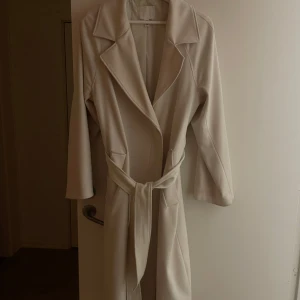 Benvit kappa med bälte - Snygg benvit kappa med bälte i midjan. Jackan har långa ärmar och en klassisk krage. Perfekt för våren och hösten, men även vintern!