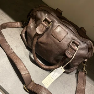 Biba boston bowling - Brun skinnväska med axelrem - Helt ny väska från BIBA med justerbar axelrem och dubbla handtag. Väskan har en dragkedja upptill. Perfekt för att bära med sig det nödvändigaste med stil.