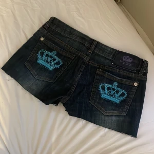 Mörkblå jeansshorts från Victoria Beckham  - Snygga mörkblå jeansshorts från Rock & Republic med coola turkosa kronbroderier på bakfickorna. Shortsen har en klassisk femficksdesign och en knappgylf. Perfekta för en avslappnad stil med en touch av lyx.