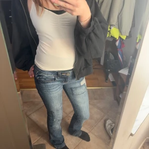 Blå jeansbyxor från Guess - Snygga blå lågmidjade jeansbyxor från Guess med en tight passform men går ut vid fötterna. Byxorna har en klassisk femficksdesign och en tydlig logga på bakfickan. Perfekta för en stilren look.