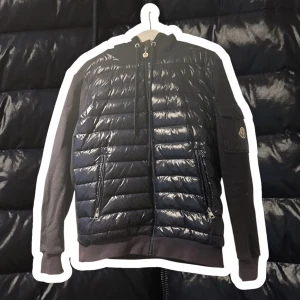 Moncler cardigan  - Snygg marinblå pufferjacka från Moncler med glansig finish och dragkedja framtill. Jackan har en huva och en praktisk ficka med knapp på ärmen. Perfekt för kyligare dagar och ger en stilren look.