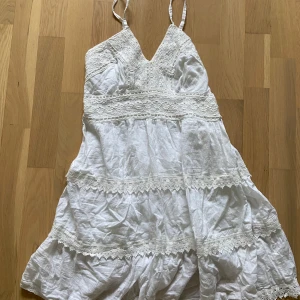 Vit klänning med spetsdetaljer - Fin vit klänning med smala axelband och v-ringad hals. Klänningen har vackra spetsdetaljer och volanger. Perfekt för sommarens varma dagar.