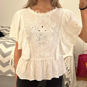 Vit blus med broderi - Charmig vit blus med vackra broderier och hålmönster framtill. Blusen har korta ärmar och en lös passform som ger en luftig känsla. Perfekt för varma dagar eller sommarens festligheter.