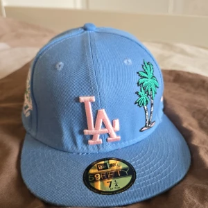 Blå keps med broderad palm och LA-logga - Snygg blå keps från New Era med broderad LA-logga i rosa och en palm i grönt. Kepsen har även ett jubileumsmärke på sidan och MLB-logga bak. Insidan är rosa med svarta detaljer. Perfekt för att ge din outfit en cool touch!