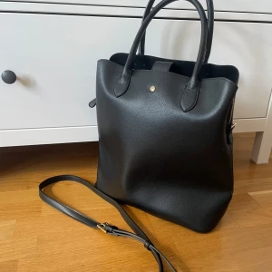 Svart väska - Snygg svart axelväska i skinn med praktisk dragkedja och avtagbar axelrem. Den är helt oanvänd och vet tyvärr inte original priset. Väskan har två handtag och en stilren design, perfekt till skolan eller jobbet. En liten knappdetalj framtill ger en extra touch. Väskan är inuti indelad i tre delar varav ena går att stänga med dragkedja. Väskan är cirka 35cm i höjd och 32cm i bredd. Pris kan diskuteras, bara fråga om fler bilder💞