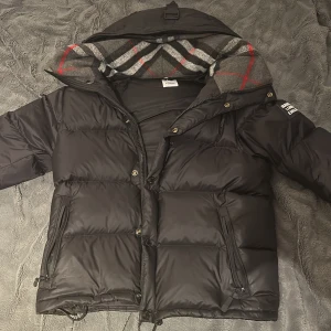 Svart pufferjacka från Burberry - Säljer en snygg svart pufferjacka från Burberry med klassiskt rutigt foder i grått och rött. Jackan har dragkedja och knappar framtill samt en huva för extra värme. Perfekt för kyliga dagar!