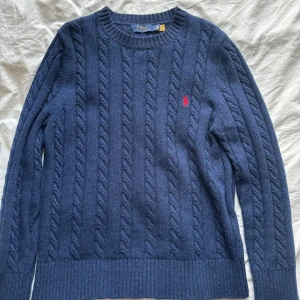 Mörkblå kabelstickad tröja från Polo Ralph Lauren - Säljer en stilren mörkblå kabelstickad tröja från Polo Ralph Lauren. Tröjan har en klassisk design med rund hals och ett rött broderat märke på bröstet. Perfekt för kyligare dagar.