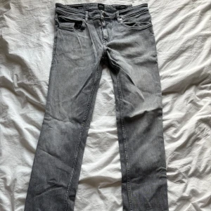 Grå jeans från Hugo Boss - Snygga grå jeans från Hugo Boss med klassisk femficksdesign och dragkedja. De har en straight passform och är perfekta för en stilren look. Passar bra till både sneakers och kängor.