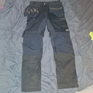 Svarta arbetsbyxor från WorkSafe - Praktiska svarta arbetsbyxor från WorkSafe med flera fickor och dragkedjor. Byxorna har förstärkta knän och är designade för att tåla tuffa arbetsmiljöer. Perfekta för dig som behöver slitstarka byxor med bra förvaring. Helt nya! 