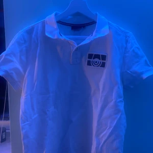 Vit polo från Sail Racing - Säljer en stilren vit polo från Sail Racing i 100% bomull. Tröjan har korta ärmar och en klassisk krage med knappar. Perfekt för en avslappnad och sportig look.