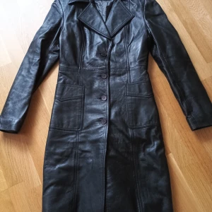 Svart skinnkappa från Roco Baroco - Snygg svart skinnkappa från Roco Baroco med klassisk design och knappar framtill. Perfekt för en stilren look. Kappan har långa ärmar och en elegant krage.