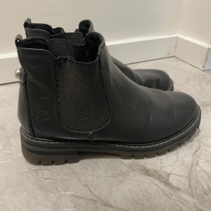 Svarta Chelsea boots från Marco Tozzi - Säljer ett par snygga svarta Chelsea boots från Marco Tozzi. De är i skinn och har en elastisk panel på sidan för enkel på- och avtagning. Skorna har en rund tå och en liten dekorativ knappdetalj på baksidan. Perfekta för höst och vinter!