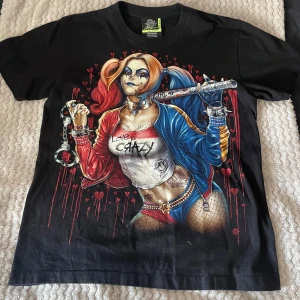 Svart t-shirt med tryck från Rock Eagle - Säljer en svart t-shirt från Rock Eagle med ett tryck av Harley quinn på både fram- och baksidan. T-shirten är i 100% bomull och har korta ärmar. Den har en 'glow in the dark' och har inga defekter. Kontakta mig om du har fler frågor 😊
