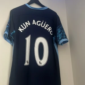Mörkblå Manchester City fotbollströja med Agüero 10 - Säljer en snygg mörkblå Manchester City fotbollströja med 'Kun Agüero' och nummer 10 på ryggen. Tröjan har korta ärmar med ett coolt mönster och är i bra skick. Perfekt för alla City-fans! 💙
