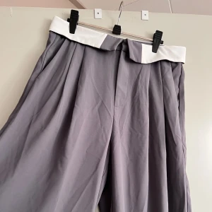 Grå vida byxor med vit linning - Säljer ett par snygga grå byxor med en vit linning. De är vida och har en elegant look, perfekt för både vardag och fest. Byxorna är i ett mjukt och bekvämt material som ger en skön passform.