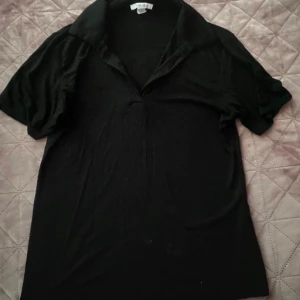 Svart kortärmad topp - Säljer en stilren svart topp med korta ärmar och en klassisk krage. Perfekt för både vardag och festliga tillfällen. Toppen är i ett mjukt och bekvämt material.