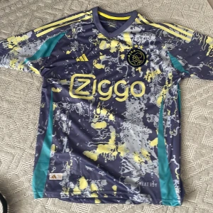 Ajax fotbollströja med mönster - Säljer en cool Ajax fotbollströja från Adidas. Den har ett unikt mönster i grått, gult och blått med korta ärmar. Perfekt för fotbollsfans som vill sticka ut på planen eller läktaren!
