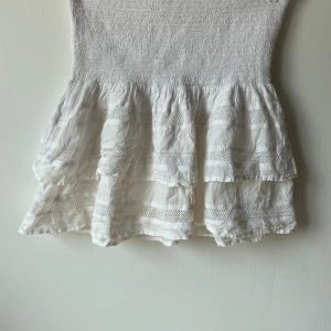 Vit kjol från Neo Noir - Säljer en superfin vit kjol från Neo Noir. Den har en söt volangdetalj och är perfekt för sommaren. Kjolen är i ett lätt och luftigt material, vilket gör den idealisk för varma dagar. Den har en elastisk midja för en bekväm passform.