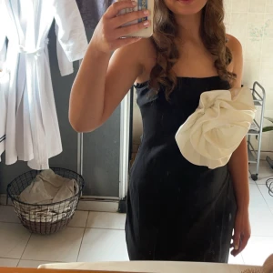 Svart axelbandslös klänning med vit detalj - Säljer en elegant svart axelbandslös klänning med en stor vit rosett-detalj framtill från Boohoo. Klänningen har en tight passform och är perfekt för festliga tillfällen. Den är i nyskick och redo att bäras på nästa stora event!