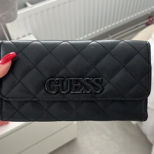Svart quiltad plånbok från Guess - Snygg svart quiltad plånbok från Guess med logotyp på framsidan. Perfekt för att hålla ordning på dina kort och kontanter. Stilren design som passar till alla tillfällen.