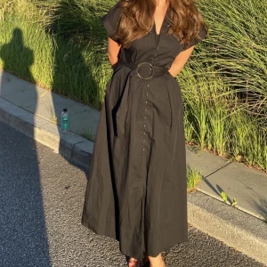 Svart klänning från Zara - Snygg svart klänning från Zara med knappar framtill och ett bälte med stor ringdetalj i midjan. Klänningen har korta ärmar och en klassisk krage. Perfekt för en stilren look.