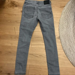 Grå jeans från Jack & Jones - Snygga grå jeans från Jack & Jones med en slim passform. De har klassisk femficksdesign och knappgylf. Perfekta för en stilren look.