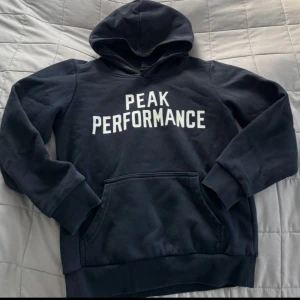 Svart/blå  hoodie från Peak Performance - Säljer en svart/blå hoodie från Peak Performance med vit text på framsidan. Den har en huva och långa ärmar. Den har även en ficka i mitten.  