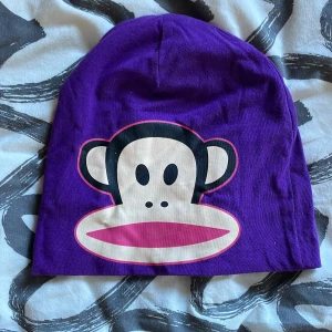Lila mössa från Paul Frank - Snygg lila mössa från Paul Frank med det ikoniska apansikte-trycket framtill. Mössan är gjord i mjukt bomullsmaterial och har en avslappnad passform. Perfekt för att ge en färgklick till din outfit!