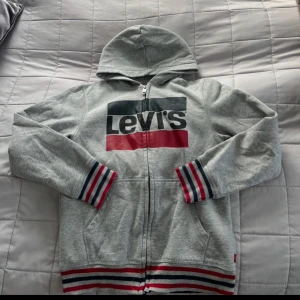Grå hoodie från Levi's - Säljer en grå hoodie från Levi's med dragkedja och huva. Tröjan har ett stort tryck med Levi's logga i svart och rött på framsidan. Ärmsluten och nederkanten har randiga detaljer i rött, vitt och blått.