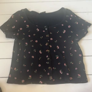 Svart blommig topp från Monki - Säljer en söt svart topp från Monki med blommigt mönster i rosa och grönt. Toppen har korta ärmar och knappar framtill. Perfekt för en avslappnad stil.