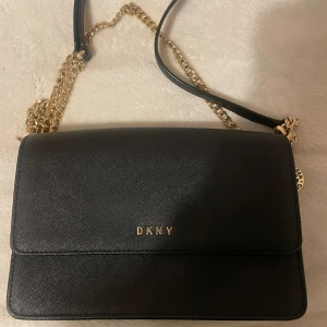 Svart axelväska från DKNY - Snygg svart axelväska från DKNY med guldfärgad kedja och logotypdetalj. Väskan har en stilren design med en praktisk storlek för vardagliga behov. Perfekt för att ge en touch av elegans till din outfit.