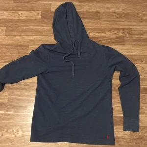 Mörkblå hoodie från Ralph Lauren - Säljer en stilren mörkgrå hoodie från Ralph Lauren med en liten röd broderad logga vid nederkanten. Tröjan har långa ärmar och en praktisk huva med snörning. Perfekt för en avslappnad look. Den är lite tunnare än vanliga hoodies.