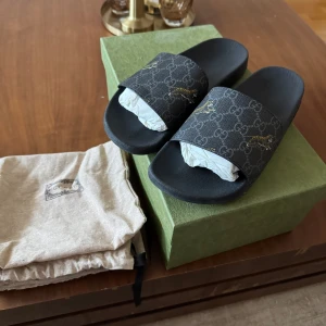 Svarta sandaler från Gucci - Snygga svarta sandaler från Gucci med ett leopard/tiger mönster. Perfekta för en stilren look. Sandalerna har en bred rem med märkets ikoniska logotyp och ett diskret mönster.