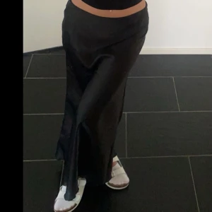 Svart långkjol i satin - Elegant svart långkjol i satin med en slits på sidan. Perfekt för en stilren look. Kjolen har en mjuk och glansig yta som ger en lyxig känsla.