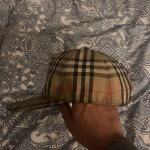 Rutig keps  - Snygg rutig keps i klassiskt beige, svart och rött mönster. Kepsen har justerbar passform baktill och är perfekt för en stilren look. Märkeslogga på baksidan ger en exklusiv touch.