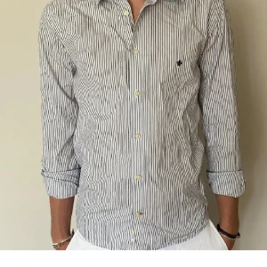 Randig skjorta med broderad logga - Snygg vit och svart randig skjorta med lång ärm och klassisk krage. Skjortan har en diskret broderad logga på bröstet och knappar framtill. Perfekt för en stilren look.