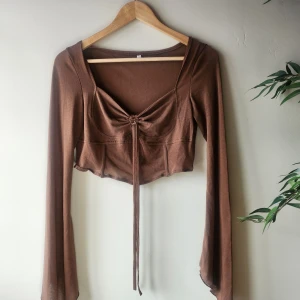 Brun croppad topp med knytning - Säljer en brun croppad topp med lång ärm och knytning framtill. Toppen har en v-ringad design och är tillverkad i ett mjukt material. Size L.