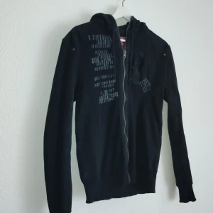 Svart hoodie från Jack & Jones - Säljer en svart hoodie från Jack & Jones med dragkedja och tryck på framsidan. Tröjan har långa ärmar och en praktisk huva. Perfekt för en avslappnad stil.