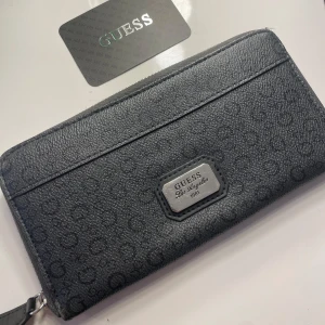 Svart plånbok från Guess - Snygg svart plånbok från Guess med diskret mönster och en metallplatta med logotyp. Plånboken har dragkedja och flera fack för kort och kontanter. Perfekt för att hålla ordning på dina värdesaker med stil. Köpte plånboken för 800kr och säljer för 450kr!