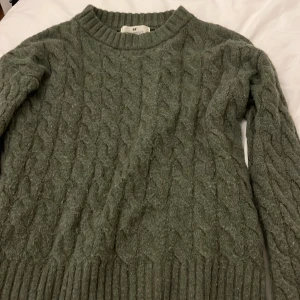 Grön stickad tröja från H&M - Säljer en mysig grön stickad tröja från H&M med ett klassiskt flätmönster. Tröjan har långa ärmar och ribbstickade muddar vid ärmslut och nederkant. Perfekt för kyliga dagar!