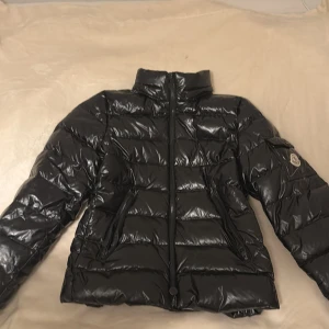 Svart dunjacka från Moncler, - Säljer en snygg svart dunjacka från Moncler. Jackan har en glansig finish och är utrustad med dragkedja framtill. Den har långa ärmar och en hög krage för extra värme. 