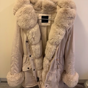 Beige parkajacka med pälsdetaljer - Snygg beige parkajacka med fluffiga pälsdetaljer på krage, ärmslut och insida. Jackan har knappar och dragkedja framtill samt praktiska fickor. Perfekt för kyliga dagar när du vill hålla dig varm och stilren.