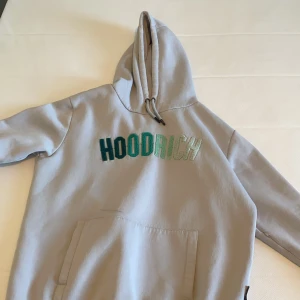 Grå hoodie från Hoodrich - Säljer en snygg grå hoodie från Hoodrich med broderad text i olika gröna nyanser på bröstet. Den har en stor ficka framtill och justerbar huva med snören. Perfekt för en avslappnad stil.
