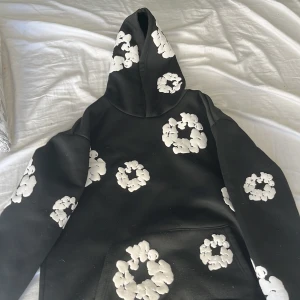 Svart hoodie från Denim Tears - Säljer en svart hoodie från Denim Tears med vita blommönster över hela plagget. Den har en klassisk huva och en stor ficka framtill. Perfekt för en unik och stilren look.