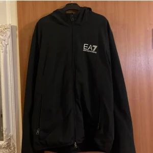 Svart jacka från Emporio Armani - Säljer en stilren svart jacka från Emporio Armani EA7. Jackan har en dragkedja framtill och en huva för extra skydd. Perfekt för en sportig look med sin minimalistiska design och diskreta logga på bröstet.Pris kan sänkas vid snabb affär.