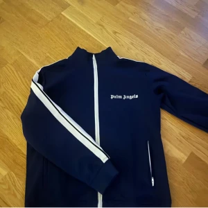 Mörkblå track jacket från Palm Angels - Säljer en snygg mörkblå track jacket från Palm Angels med vita detaljer. Jackan har dragkedja framtill och vita ränder längs ärmarna. Perfekt för en sportig look!
