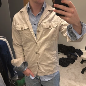 Beige övershirt från East West - Säljer en stilren beige overshirt från East West. Kavajen har knappar framtill och två bröstfickor med lock. Perfekt för en avslappnad men ändå uppklädd look. Passar bra över en skjorta för en snygg stil. Nypris på denna är runt 1300kr. Skriv vid funderingar!